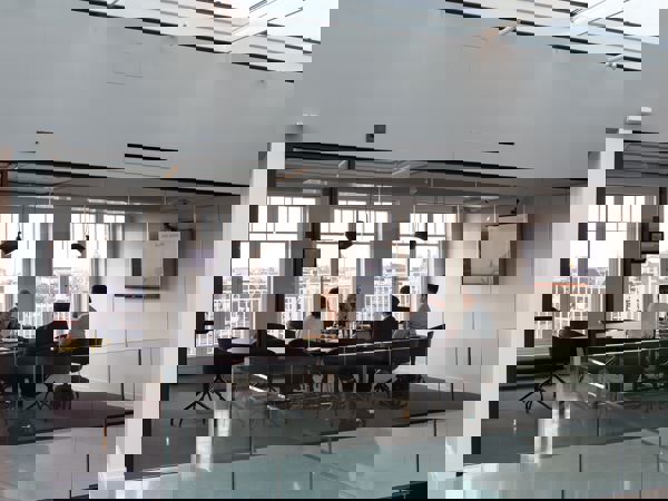 En grupp i ett modernt mötesrum med glasväggar och stor skärm, engagerade i diskussion med utsikt över staden genom fönstren.