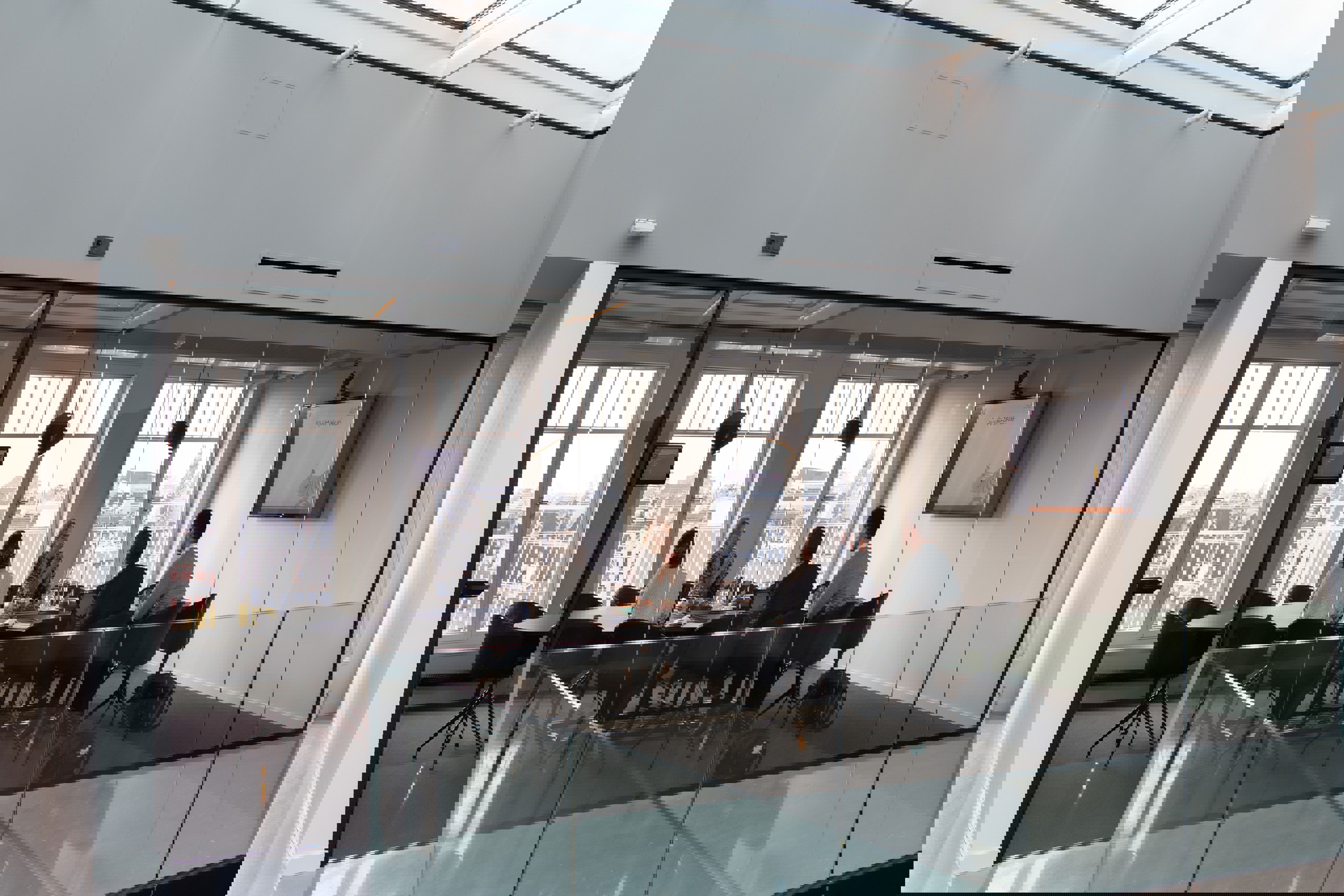 En grupp i ett modernt mötesrum med glasväggar och stor skärm, engagerade i diskussion med utsikt över staden genom fönstren.