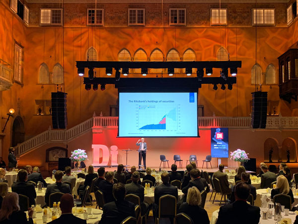 Stor konferens i elegant sal med scenpresentation om ekonomiska trender, omgiven av publiken sittande vid runda bord.