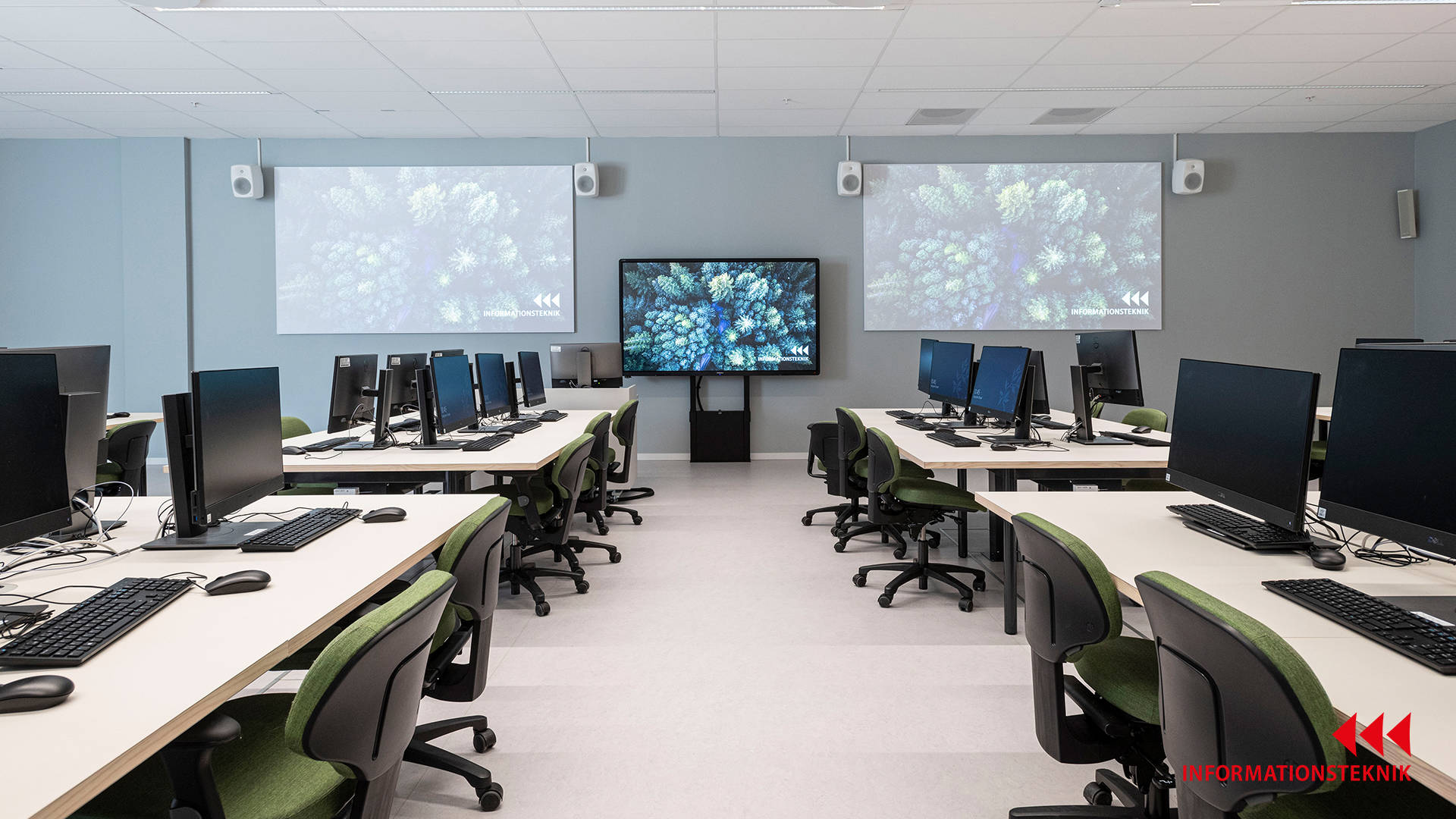 Moderna datorarbetsstationer i klassrum med projektorer och skärm för presentationer och interaktiva lektioner.