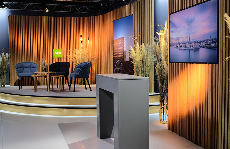 En studio med elegant studiodekor, inkluderande träpaneler, blå fåtöljer och skärmar som visar stadsbilder, upphöjd scen och belysning.