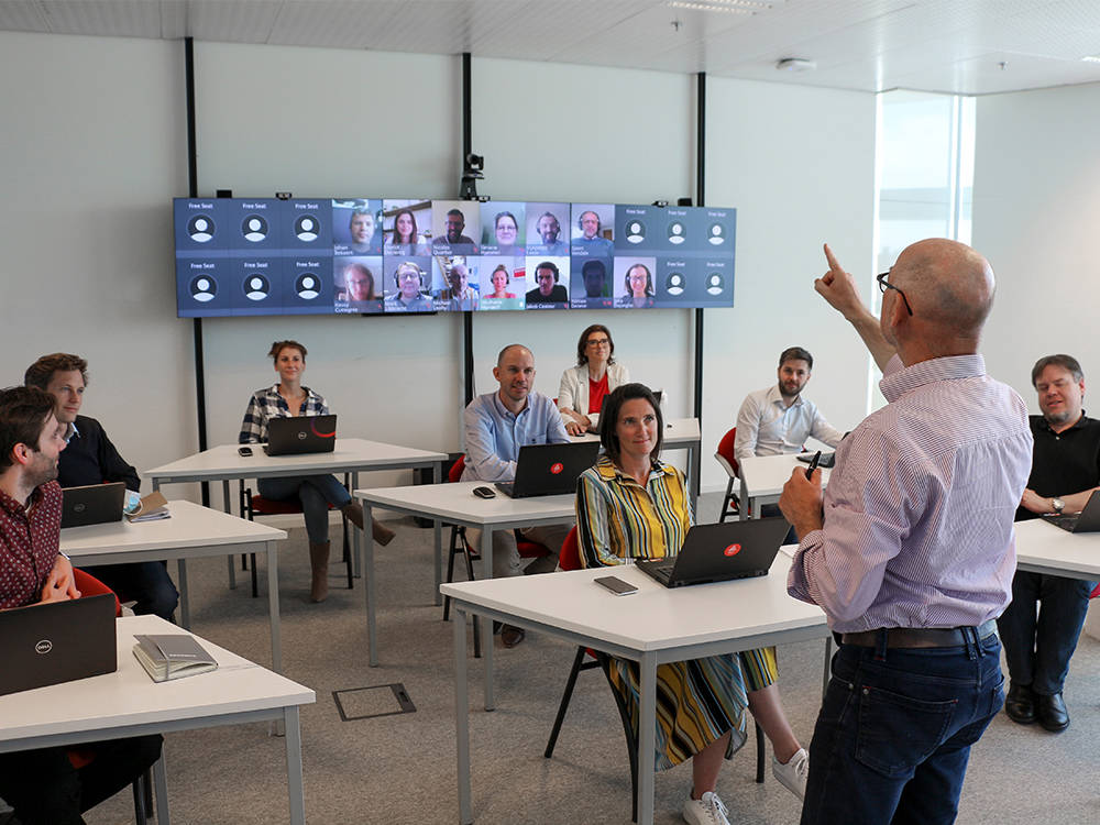 Digitalt klassrum med deltagare på plats och på skärm via videolänk, där en lärare föreläser och interagerar med båda grupper.