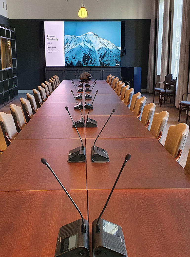 Konferensrum med långbord, mikrofoner och stor skärm för videomöte. Stolar i rad och bergsbild visas på skärmen.