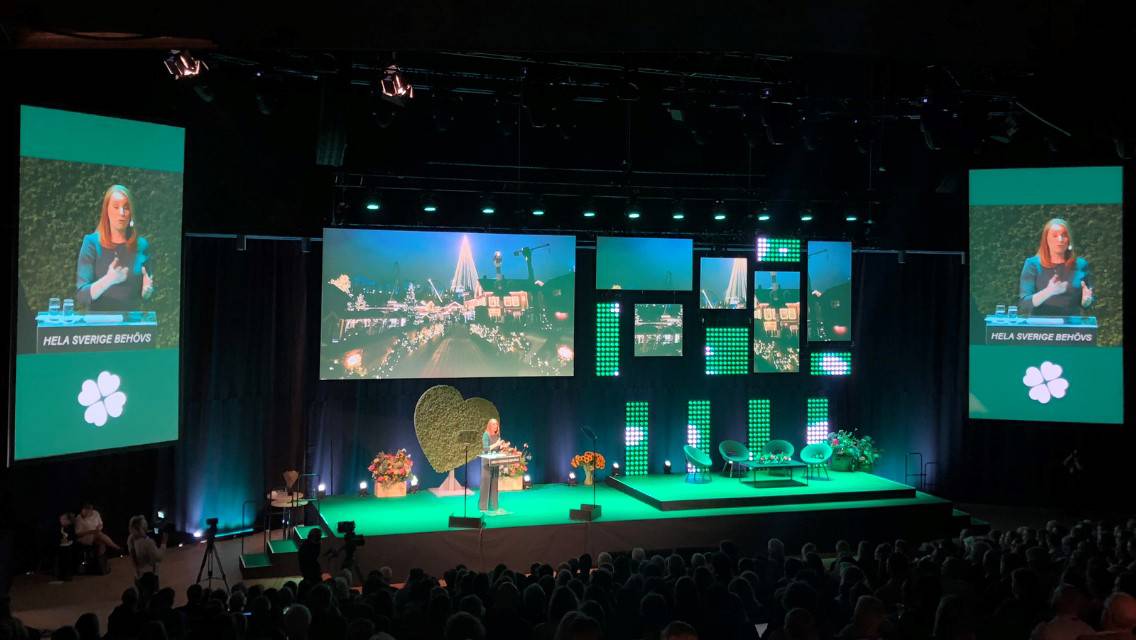 Konferens med talare på scen och flera skärmar som visar bilder, omgiven av grön belysning och publik i bakgrunden.