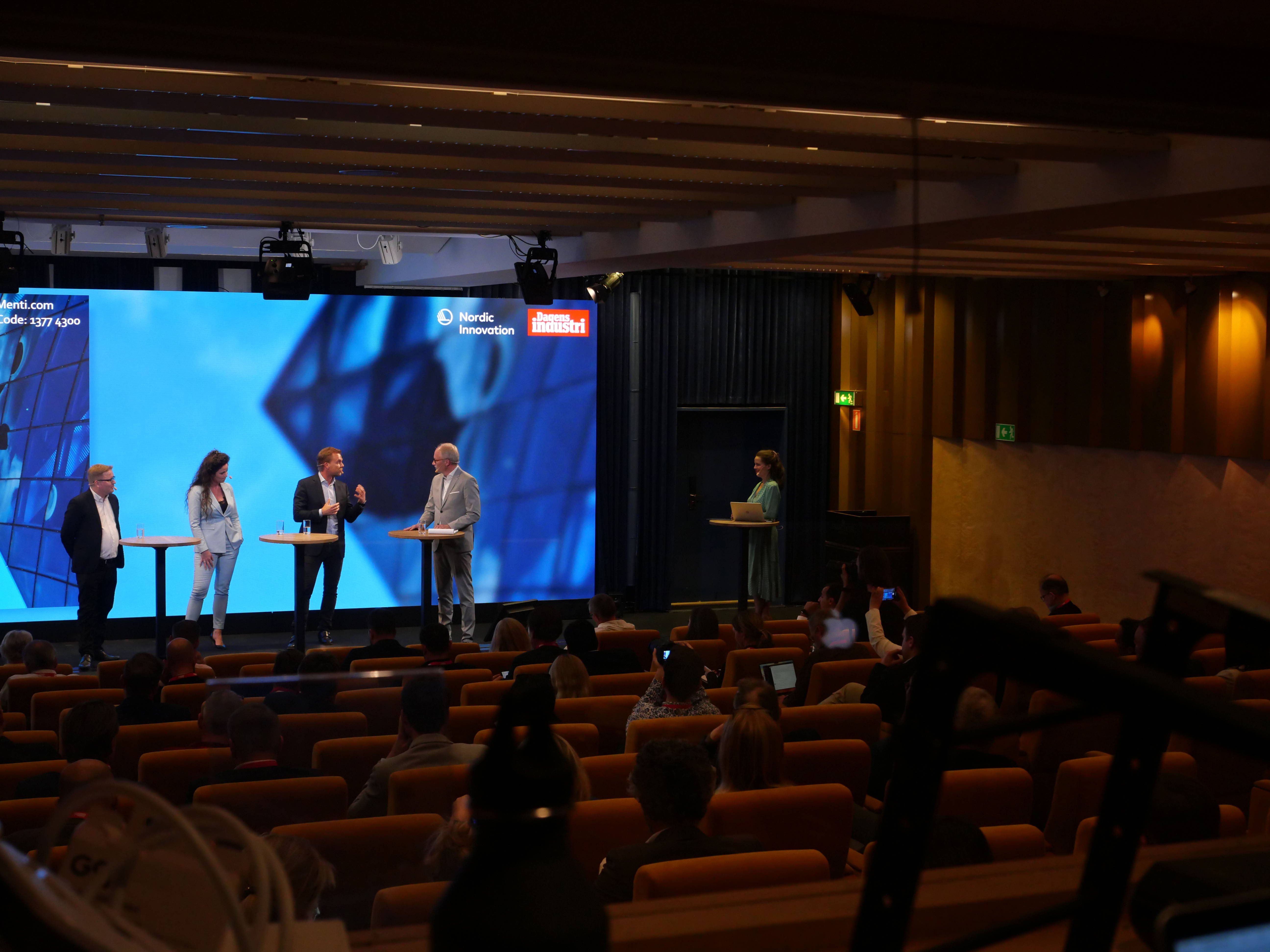 Paneldiskussion på en scen, med publik i salongen och stor skärm som bakgrund för presentationen.