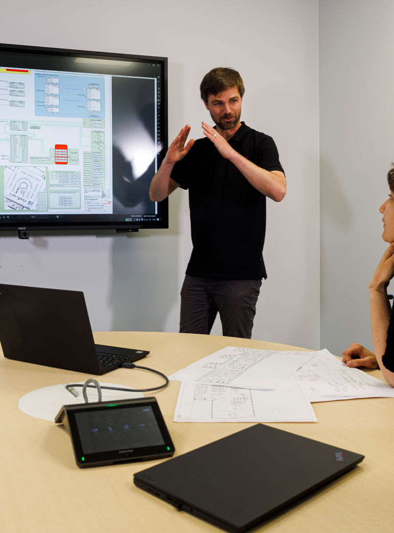 En grupp diskuterar rumstypslösningar vid ett möte, med en presentation på en stor skärm och laptopar på bordet.