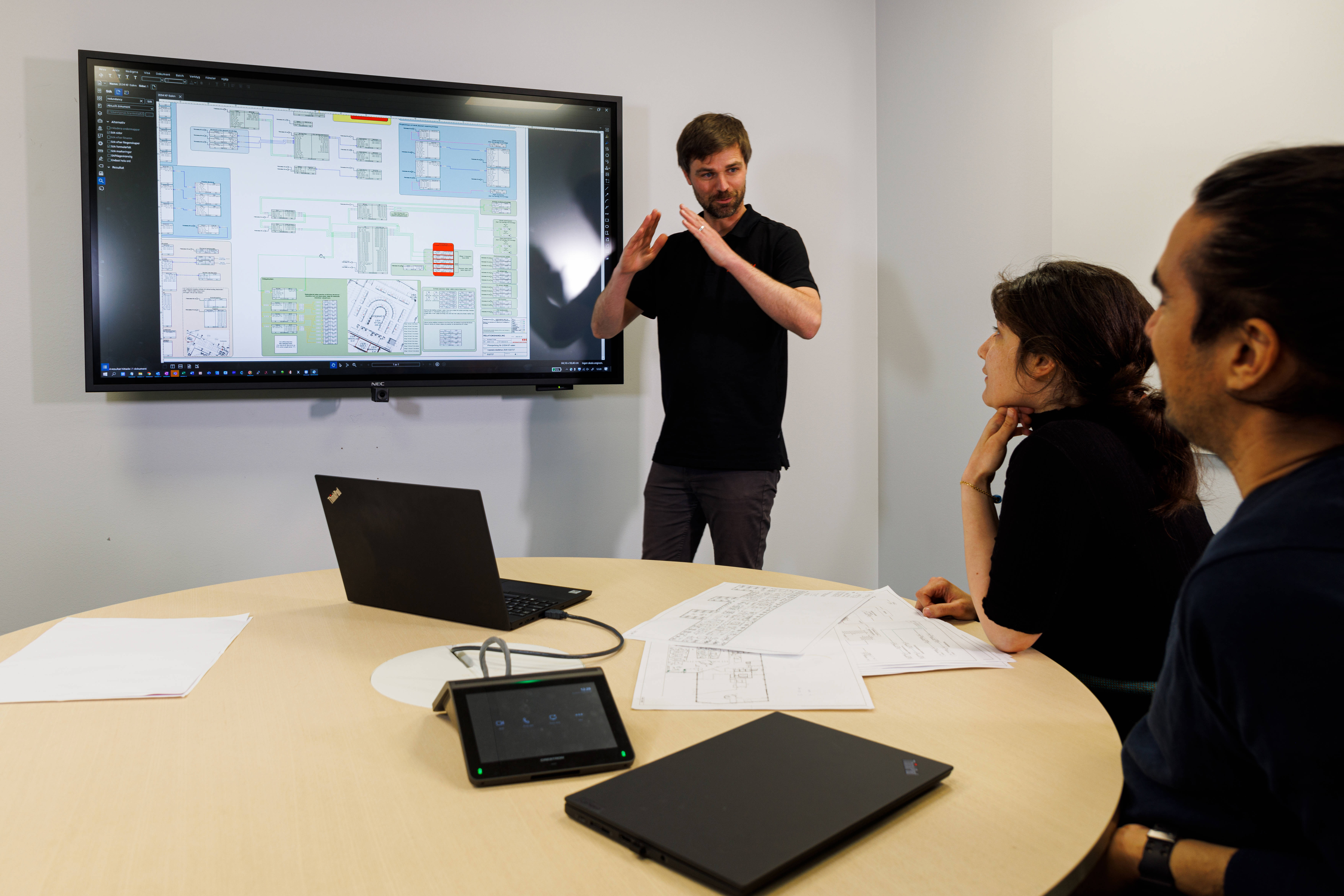 Diskussion i mötesrum med skärm och teknisk presentation; deltagare engagerade i genomgång av komplexa planer och diagram